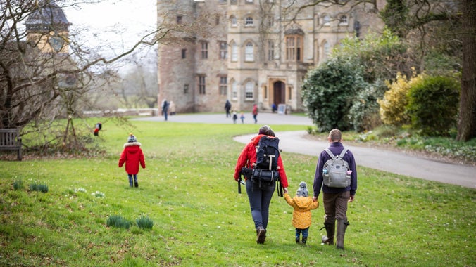 two adults in rain coats holding a small child's hands as they walk towards the castle along the grass.